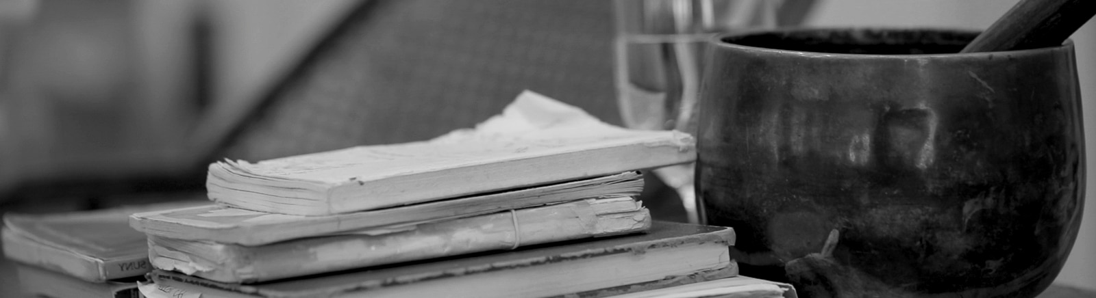 A black and white photograph with a dark colored singing bowl sitting to the right and a stack of weathered old books sits to the left while a glass of water is in the background.