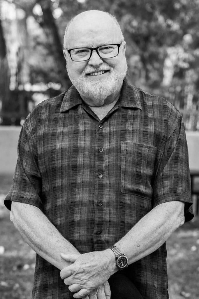A black and white photo of Richard Rohr in a plaid shirt wearing black-rimmed glasses standing in the backyard of CAC.
