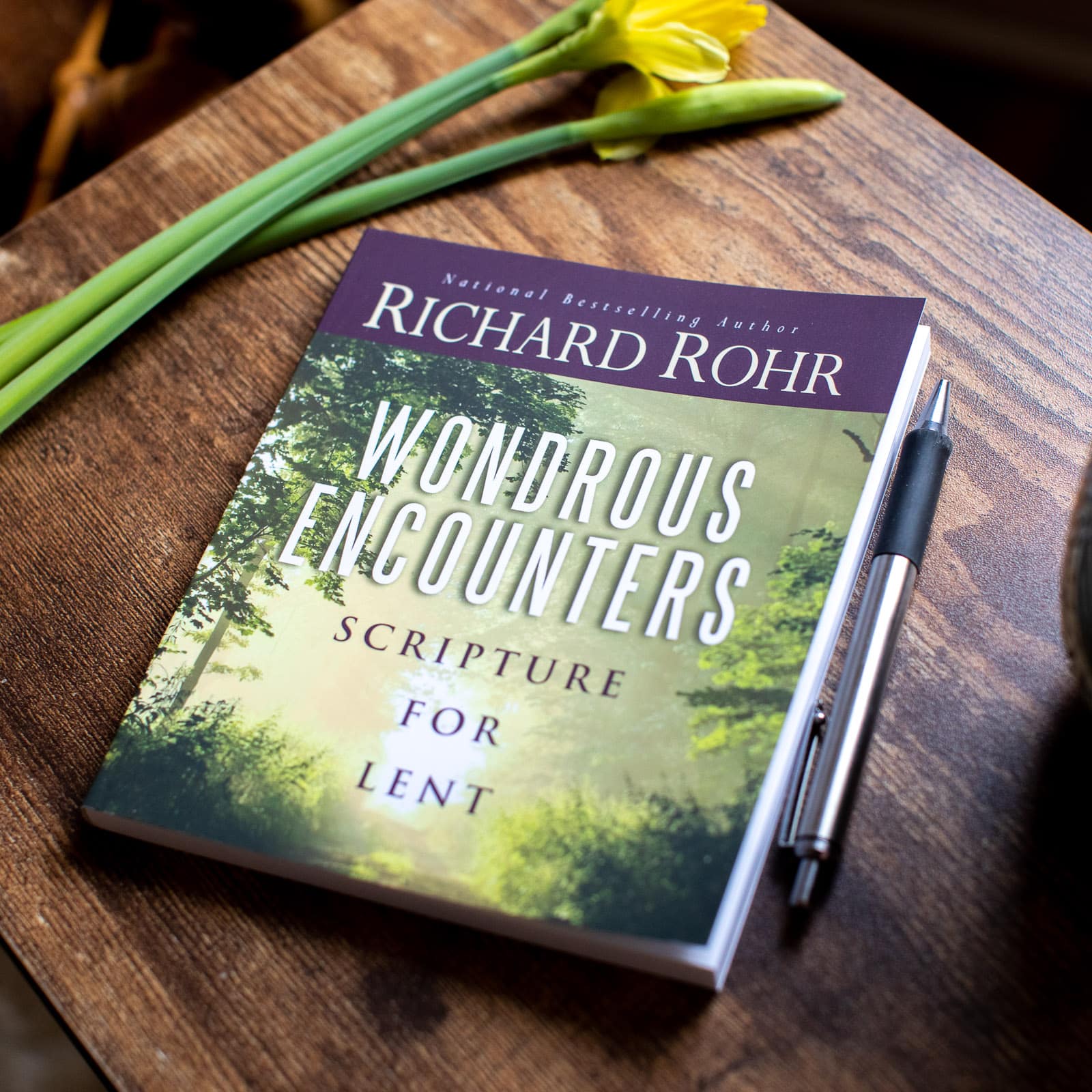 Book "Wondrous Encounters: Scripture for Lent" by Richard Rohr on a table with a pen and daffodil, ideal for spiritual reading during Lent.