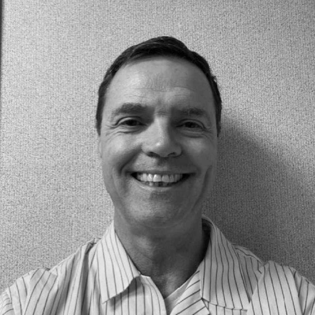 Black and white photo of Joe Lonergan wearing a stripped button up smiling in front of a wall.