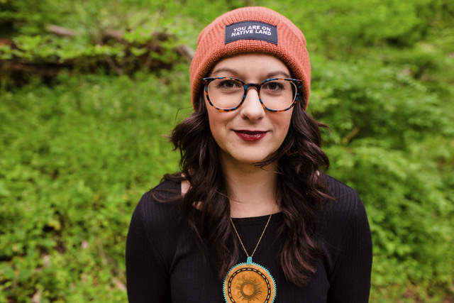 Woman wearing an orange beanie and glasses standing in a lush green forest, showcasing a natural setting and stylish fall fashion.