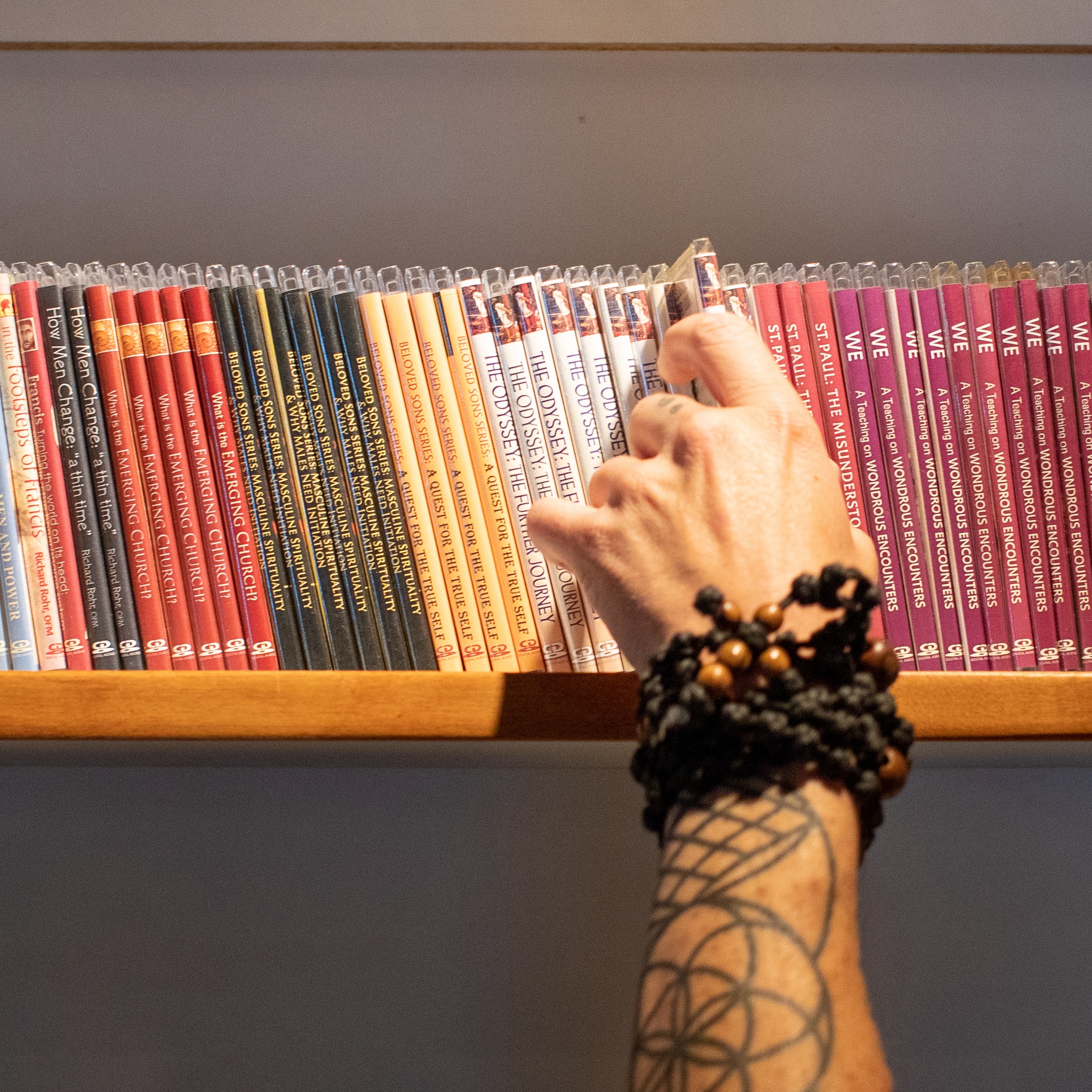 A tattooed hand selects a book from an organized shelf with various covers showcasing different literary themes.