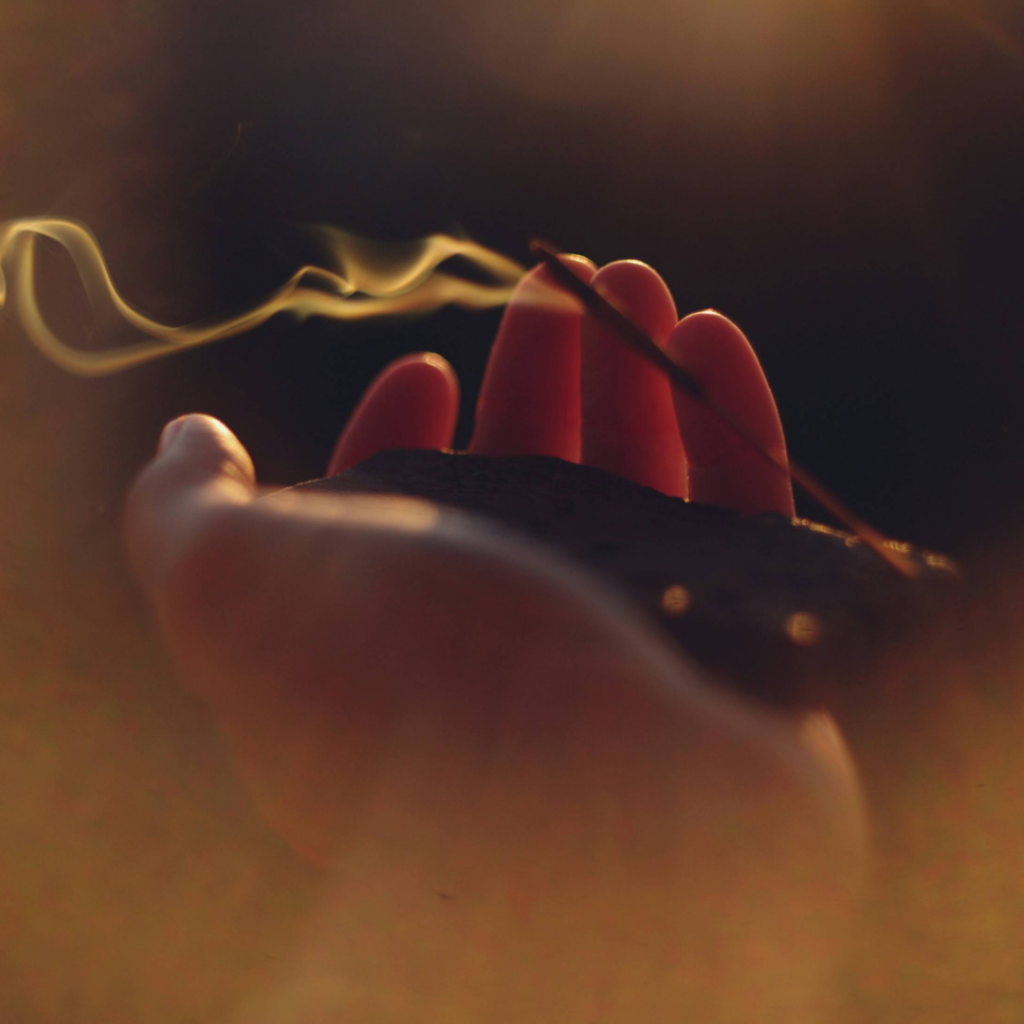 Close-up of a hand holding a burning incense stick, smoke curling gently upwards against a dark background, evoking calmness and relaxation.