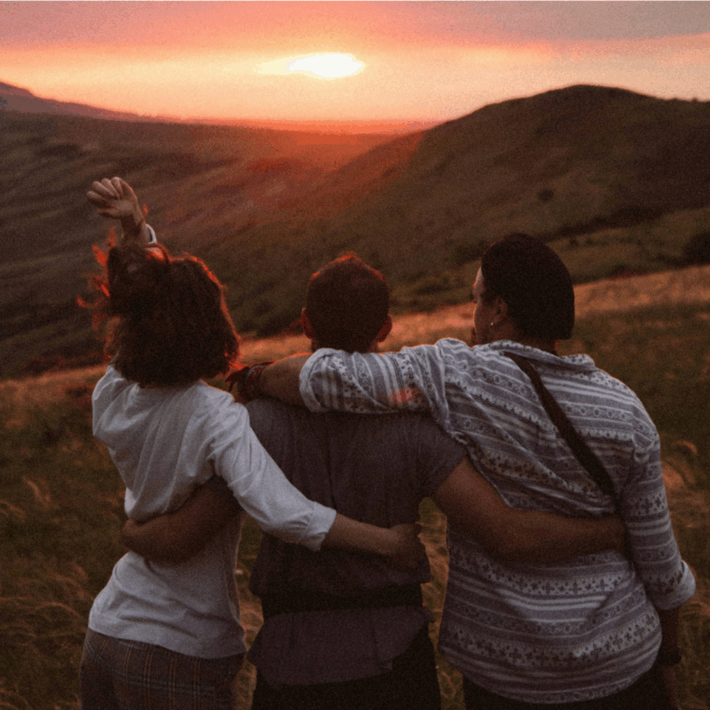 Three friends embrace on a grassy hilltop, watching a vibrant sunset over the distant mountains, enjoying nature and camaraderie together.