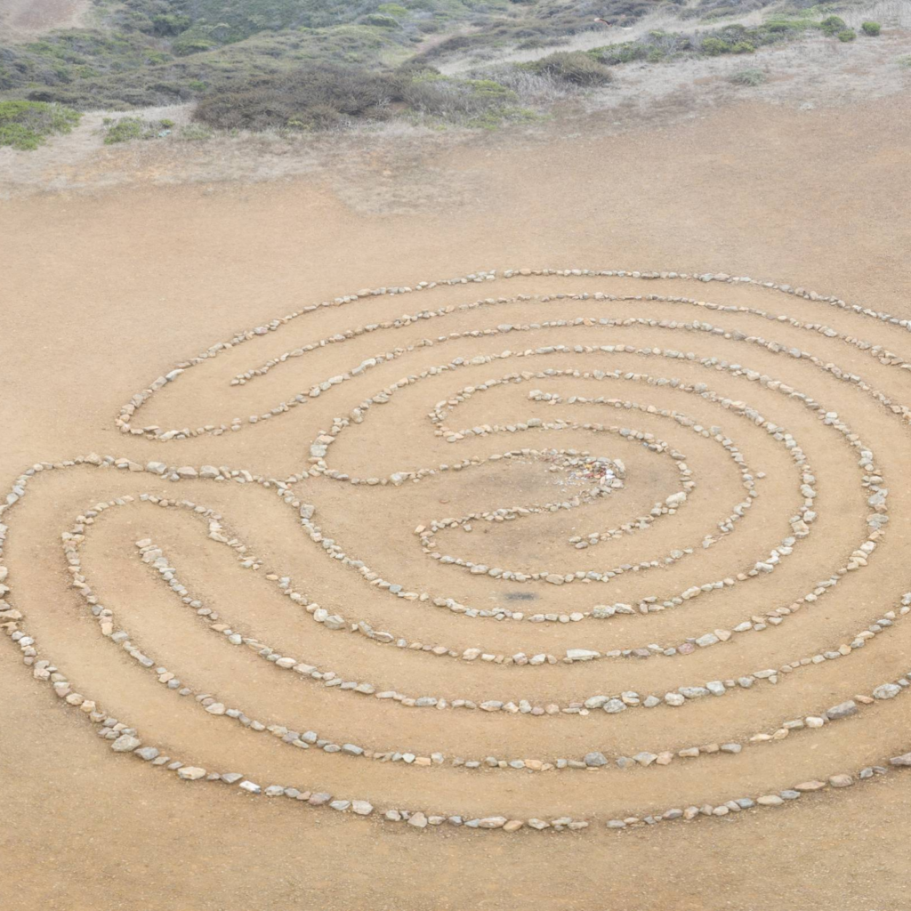 An aerial view of a large, intricate labyrinth made of small, light-colored stones arranged in a spiral pattern on a sandy, earthy ground. The labyrinth is surrounded by sparse, dry vegetation and hills in the distance, creating a serene and natural landscape.