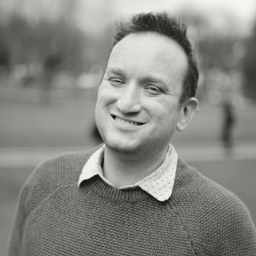 Smiling man in a cozy sweater stands outdoors, showcasing a cheerful expression in a blurred park background.
