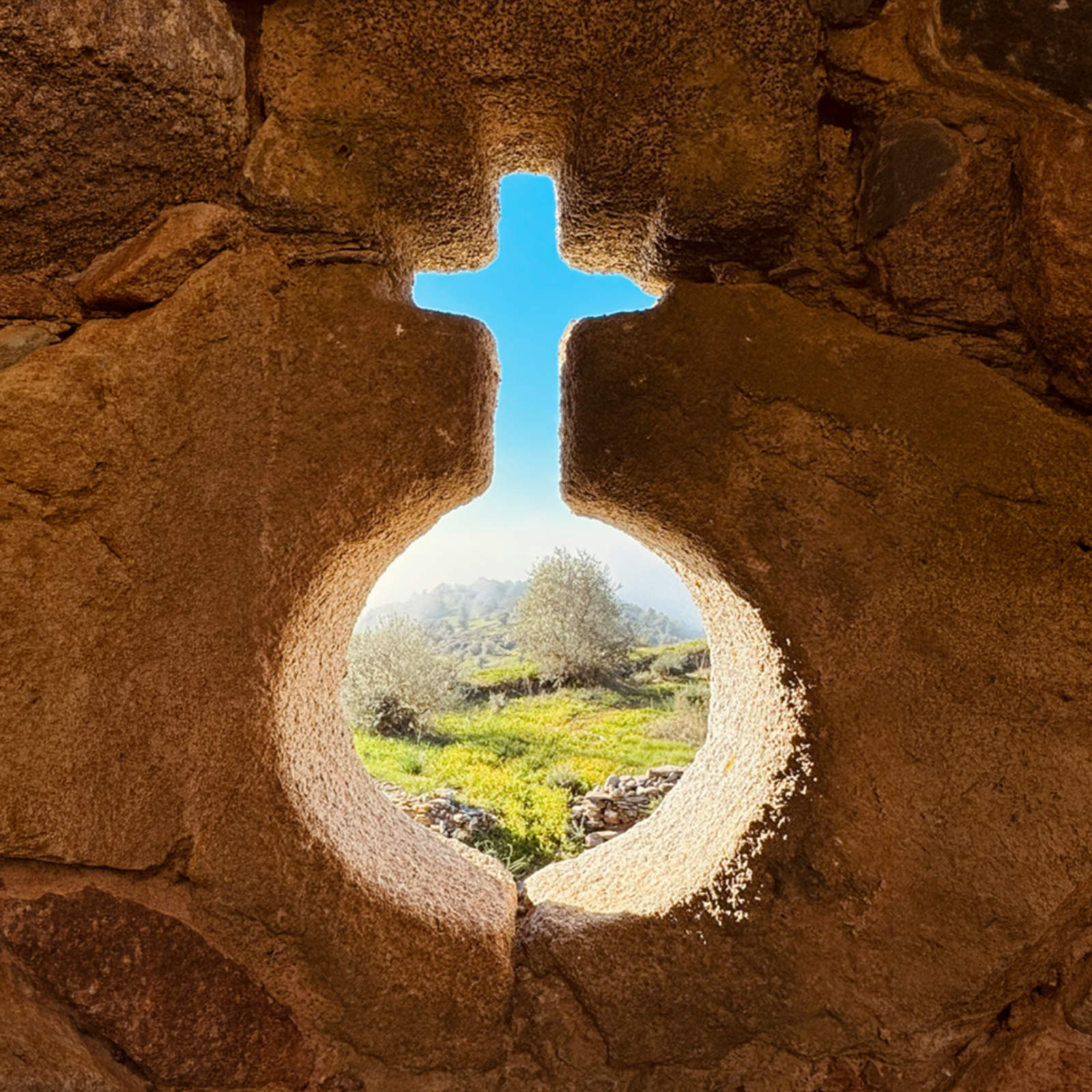 View through a stone window shaped like a cross, showcasing a serene landscape with olive trees and blue sky.