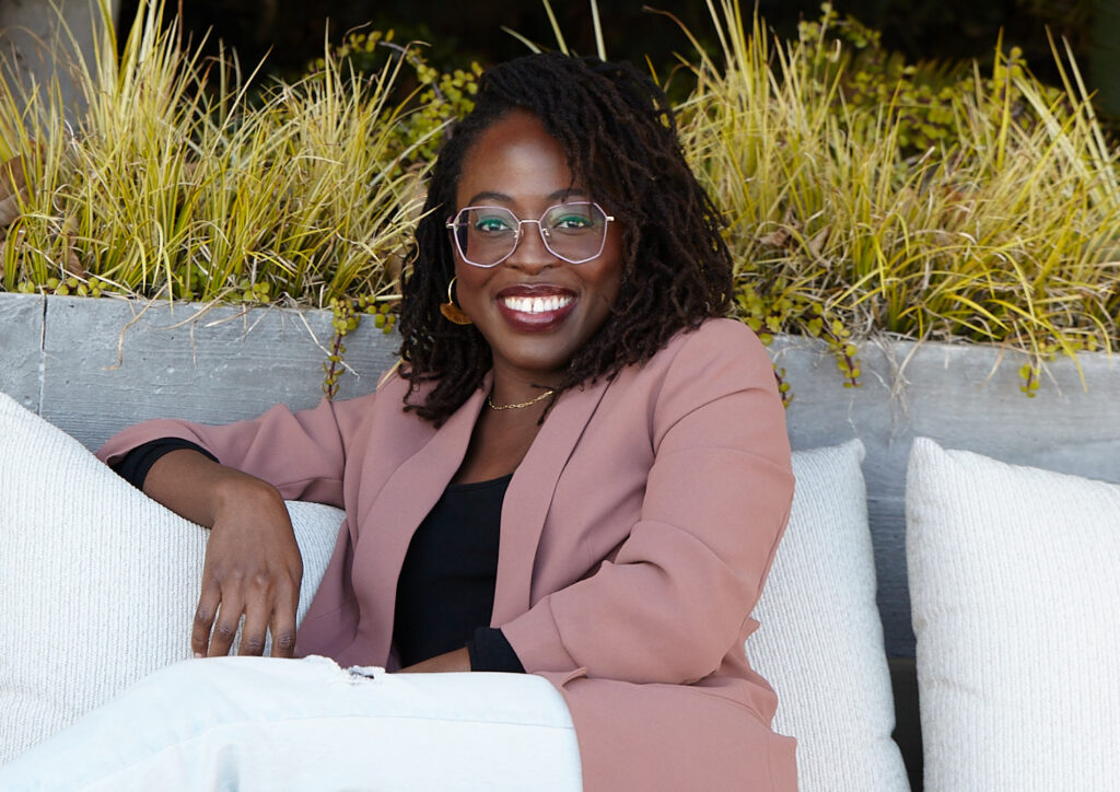 Author and Enneagram expert Chichi Agorom smiling in a rose colored blazer as she sits on a cushioned bench.